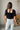 Full body/back view of model wearing the Stand Alone Top which features a black material, a scoop neckline with a ruffle hem, a short puffy sleeve, a smocked front, a flowy bottom he, a cropped fit, a smocked back, and a tie back.