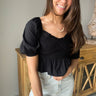 Front view of model wearing the Stand Alone Top which features a black material, a scoop neckline with a ruffle hem, a short puffy sleeve, a smocked front, a flowy bottom he, a cropped fit, a smocked back, and a tie back.