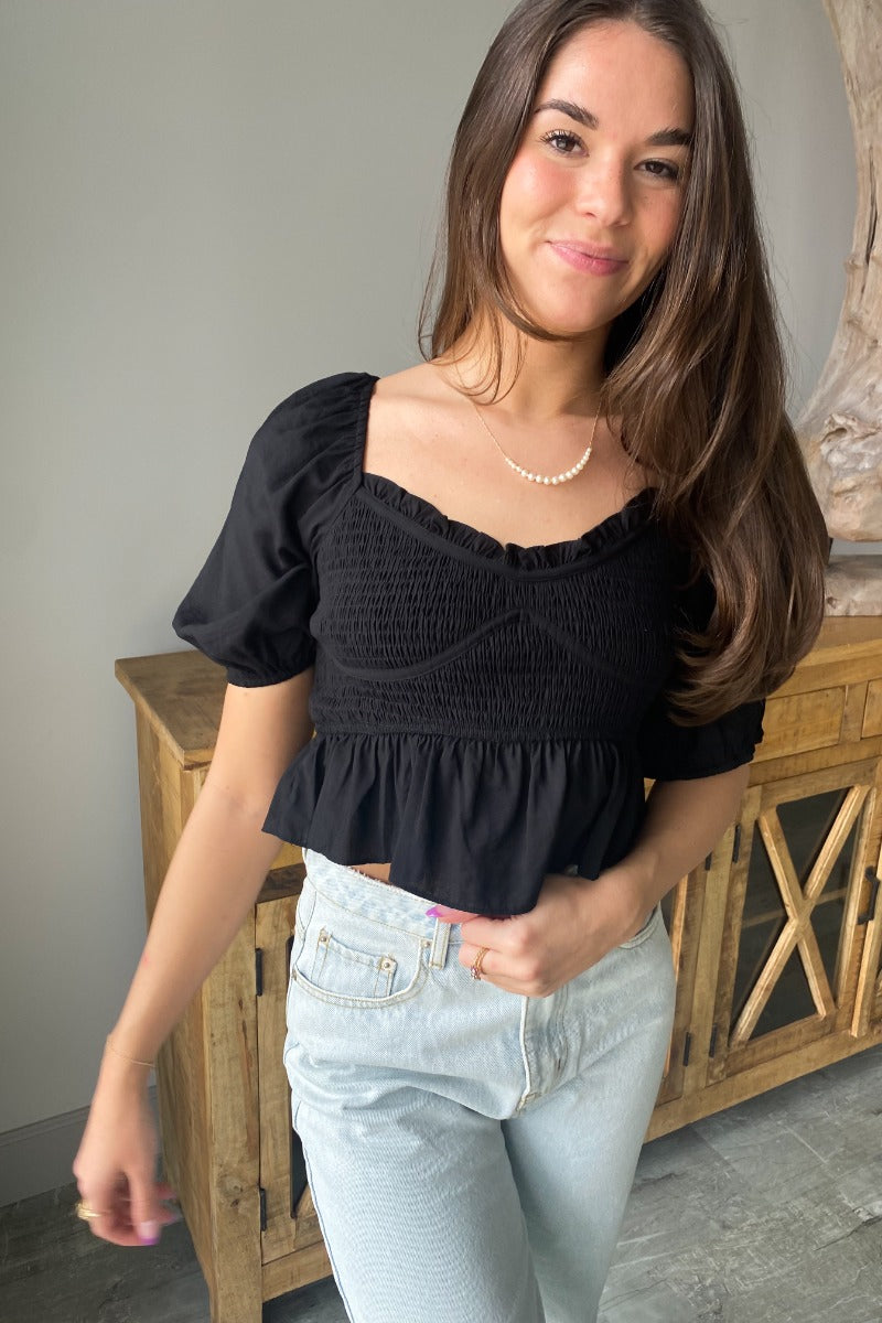 Close up view of model wearing the Stand Alone Top which features a black material, a scoop neckline with a ruffle hem, a short puffy sleeve, a smocked front, a flowy bottom he, a cropped fit, a smocked back, and a tie back.