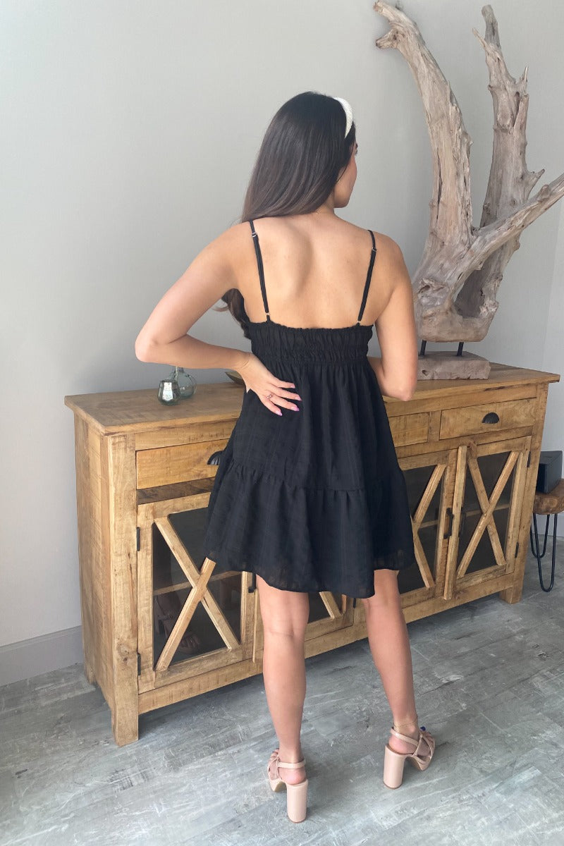 Full body/back view of model wearing the Better Than Basic Dress which features black fabric with a monochromatic plaid design, ruffle trim, a mini length hem, black lining, a smocked elastic high waistband, a low v-neckline, an open back, and adjustable 