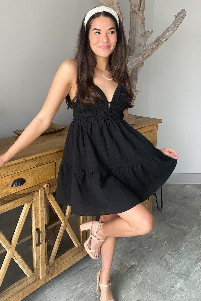 Full body view of model wearing the Better Than Basic Dress which features black fabric with a monochromatic plaid design, ruffle trim, a mini length hem, black lining, a smocked elastic high waistband, a low v-neckline, an open back, and adjustable spagh