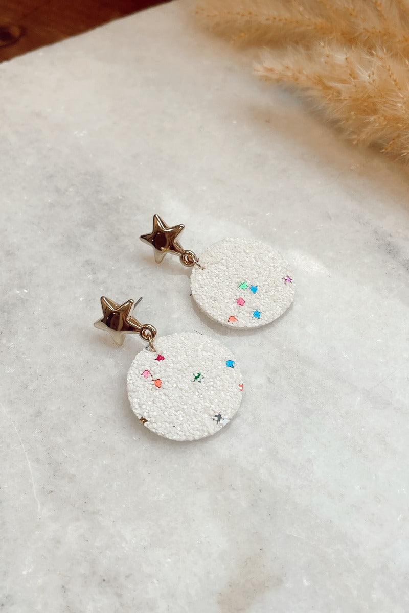 Ariel view of the Stars In The City Earring which features white, speckled circle shaped earring with multi color stars and attached to gold star clasps.