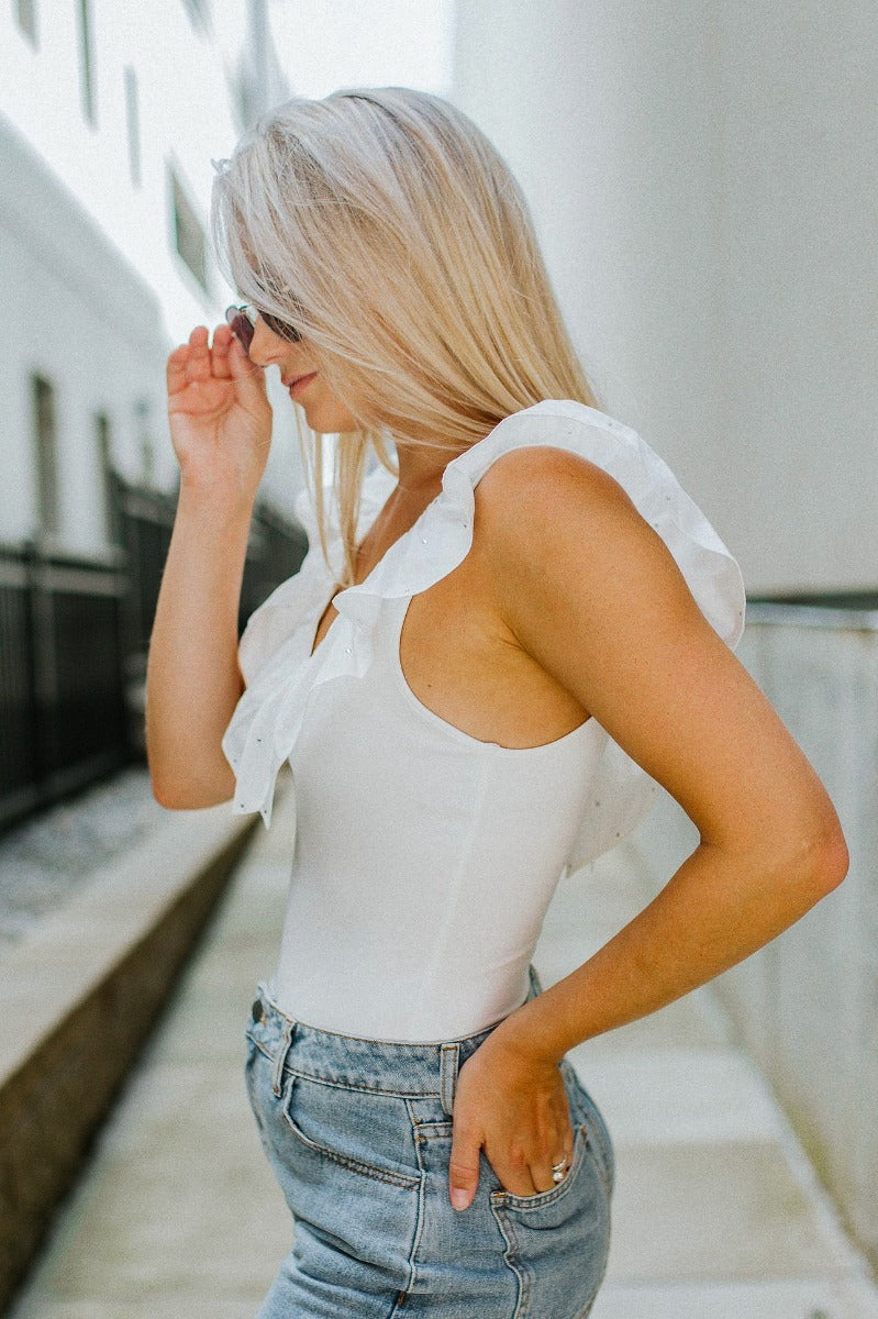 side view of model wearing the Keep Shining Rhinestone Bodysuit that has white knit fabric, a v-neck, ruffle straps with rhinestones, and a thong bottom with snap closures
