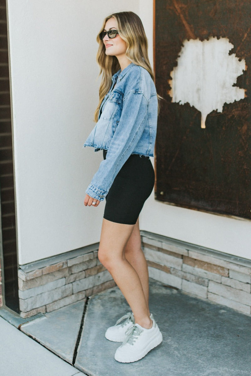 Full body side view of model wearing the Over it Cropped Denim Jacket that has blue denim fabric, a cropped waist with a raw hem, a button up front, pockets, a collared neck, and long sleeves.