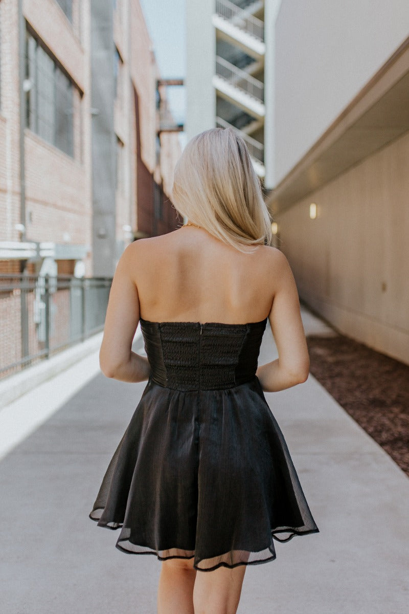 Back view of model wearing the Love Of Mine Dress that has black sheer fabric, mini length, black lining, a strapless sweetheart neckline with pleating, and a smocked back.