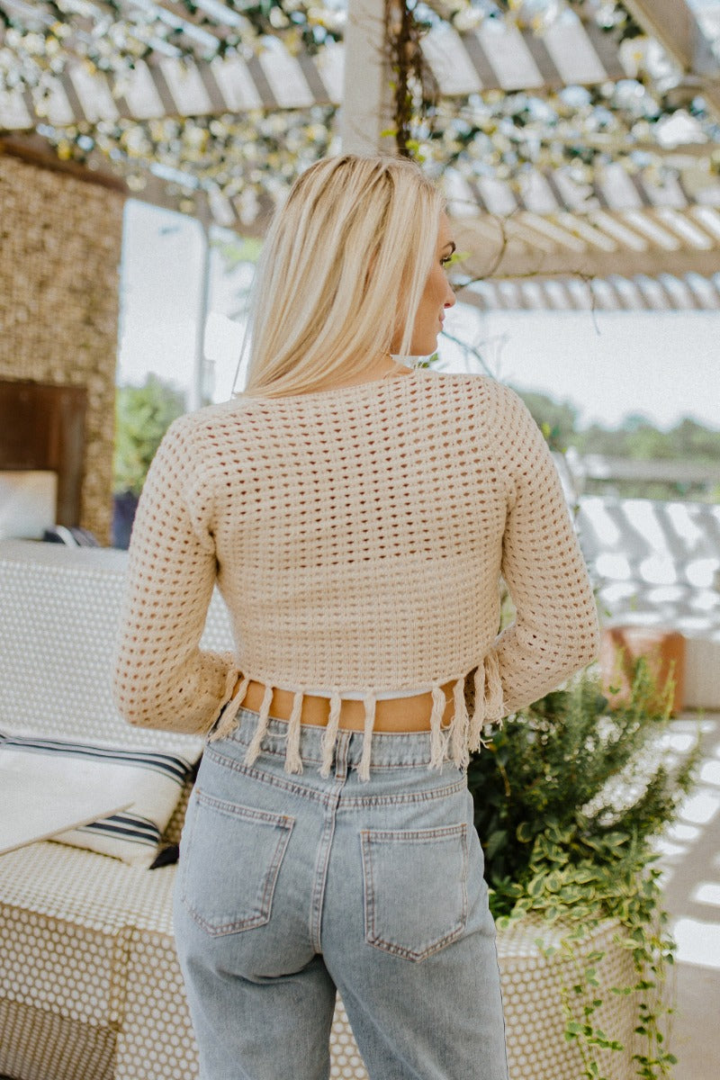 Back view of model wearing the Heading West Open-Knit Top that has taupe open knit fabric, a cropped waist with tassel hem, a square neckline and long sleeves