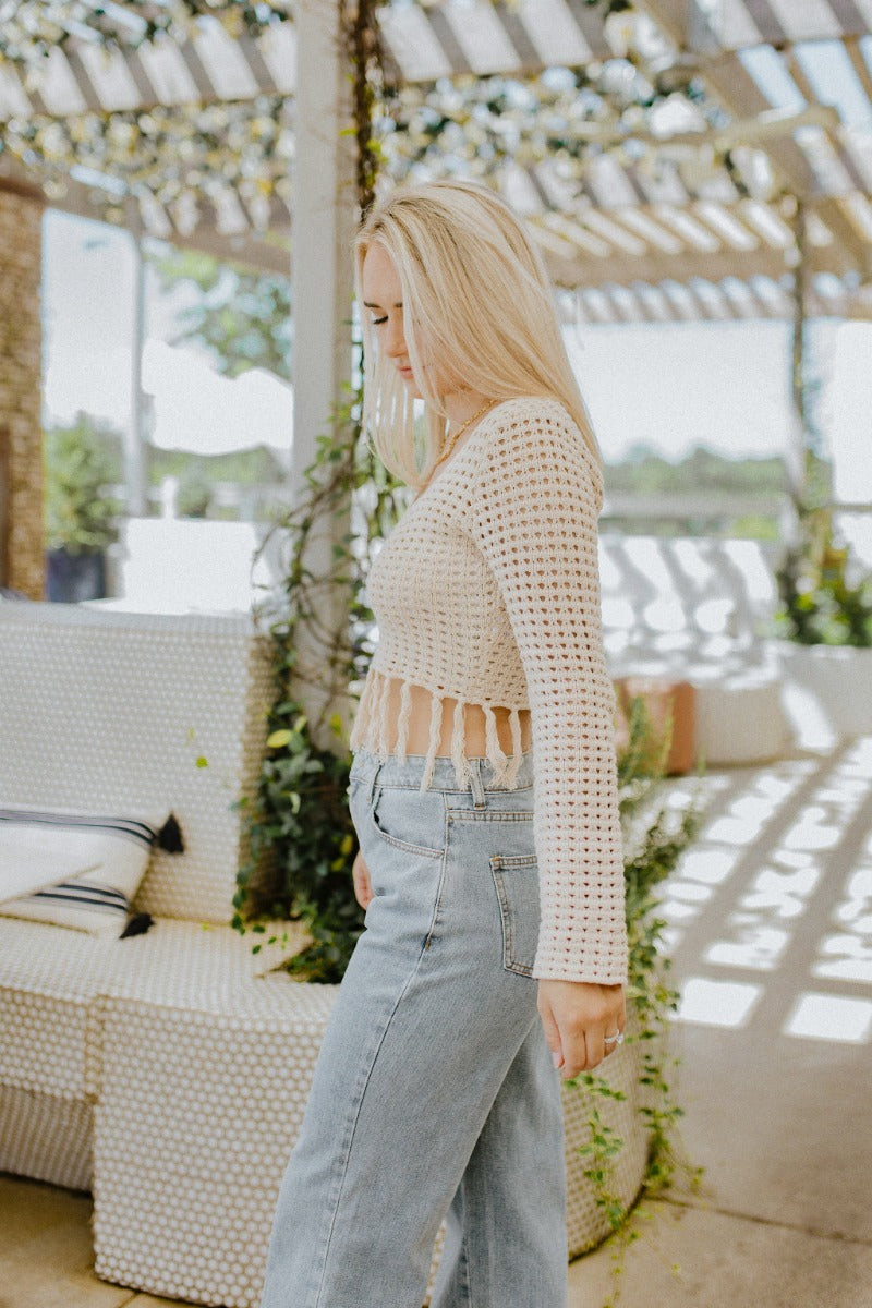 Side view of model wearing the Heading West Open-Knit Top that has taupe open knit fabric, a cropped waist with tassel hem, a square neckline and long sleeves