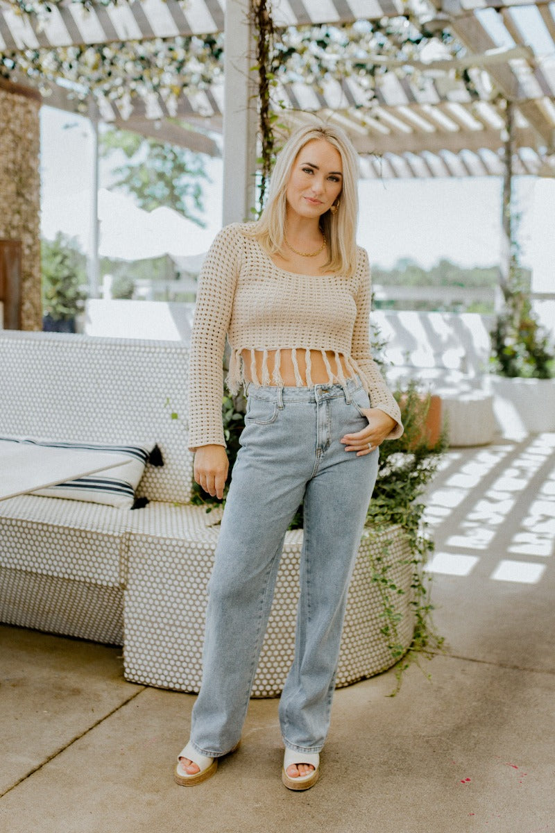 Front view of model wearing the Heading West Open-Knit Top that has taupe open knit fabric, a cropped waist with tassel hem, a square neckline and long sleeves