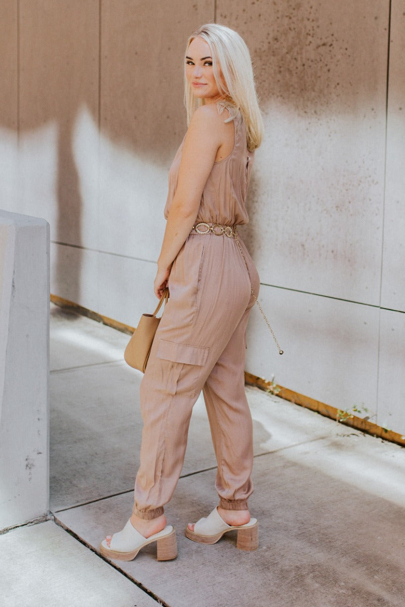 Full side view of model wearing the Roxanne Satin Cargo Jumpsuit that has dark taupe satin fabric, tapered legs with elastic ankles, cargo pockets, an elastic waist, a v-neck, a sleeveless body, and a button up closure in the back.