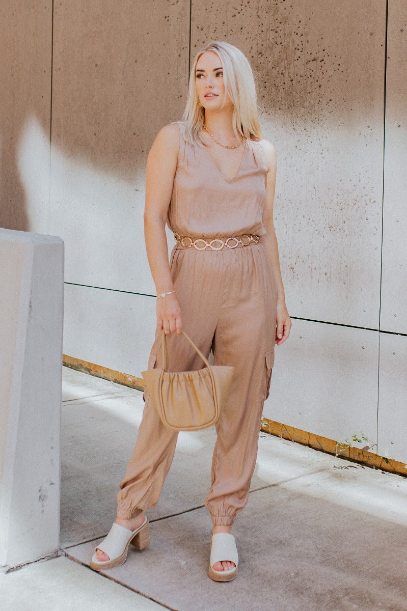 Full front view of model wearing the Roxanne Satin Cargo Jumpsuit that has dark taupe satin fabric, tapered legs with elastic ankles, cargo pockets, an elastic waist, a v-neck, a sleeveless body, and a button up closure in the back.