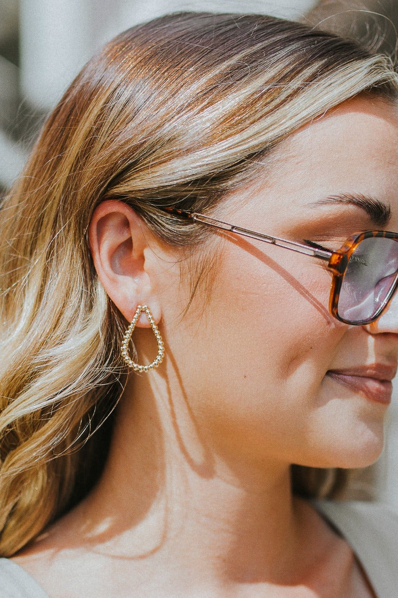 Side view of model wearing the Nothing Is Forever Earrings which features small, teardrop shaped hoops with small gold beads.