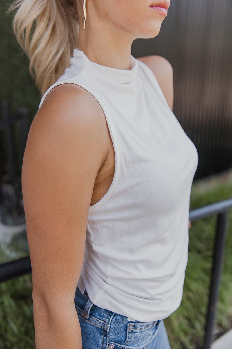 Close side view of model wearing the True Story Mock Neck Tank in White that has white knit fabric, a high neckline, and a sleeveless design.