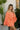 Side view of model wearing the Take Me To The Tropics Romper that has coral fabric, an elastic waist, a tie around the waist, an off-the-shoulder neck, and 3/4 flare sleeves with ruffle trim.