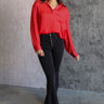 Full body front view of model wearing the Julianna Red Satin Long Sleeve Top that has red satin fabric, a high-low hem, a chest pocket, a notched collar neck, and long sleeves with buttoned cuffs.