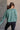 back view of model wearing the Nicole Green Cropped Long Sleeve Sweatshirt that has gray-green cotton fabric, a cropped waist, a round neckline and long sleeves.