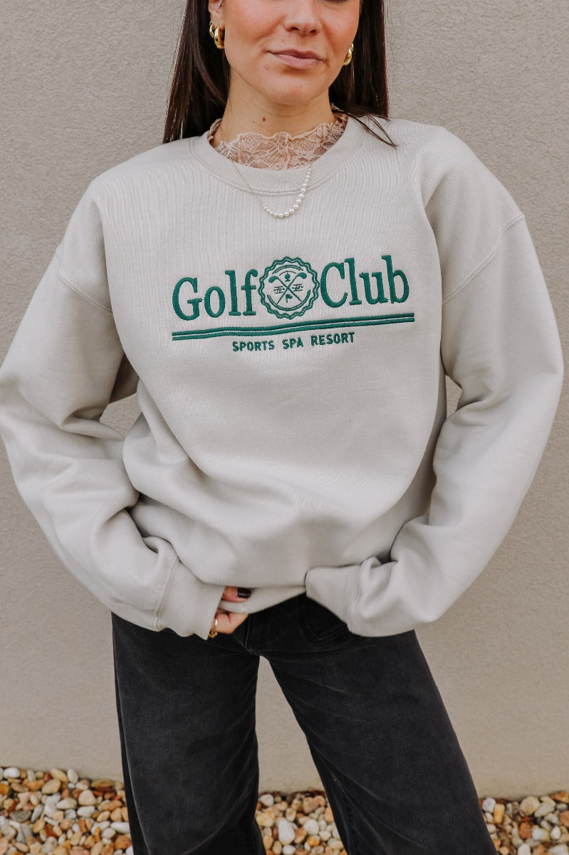 Close up view of model wearing the Golf Club Beige & Green Sweatshirt which features beige knit fabric, a round neckline, and long sleeves with ribbed cuffs. Embroidered graphic says "Golf Club" "Sports Spa Resort" with golf clubs symbol in green threadin