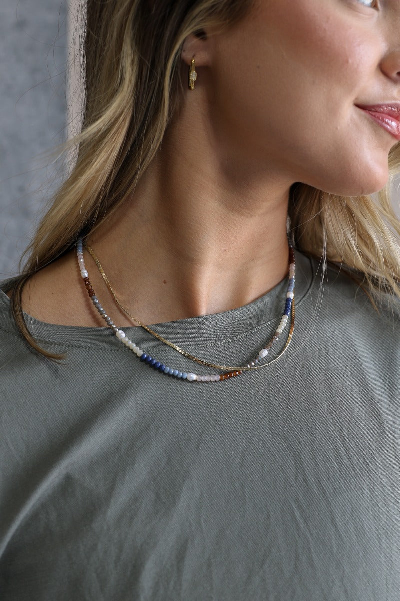 Image of model wearing the Josephine Beaded Gold Layered Necklace that features two layeres: one flat gold chain and one beaded strand with navy, light blue, brown, blush, and pearl beads.