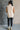 Full body back view of model wearing the Olivia Beige Cable Knit Turtleneck Sleeveless Sweater which features beige cable knit fabric, high low hem, turtleneck neckline and sleeveless.