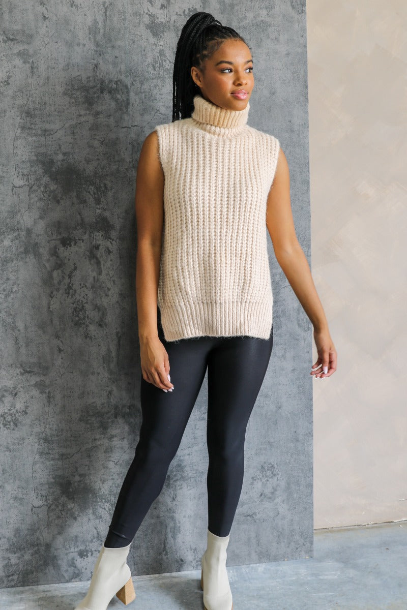 Full body view of model wearing the Olivia Beige Cable Knit Turtleneck Sleeveless Sweater which features beige cable knit fabric, high low hem, turtleneck neckline and sleeveless.