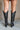 Back view of model wearing the Arcadia Black Western Boots that feature black faux leather, cream western stitching, and black soles with 3 inch heels.