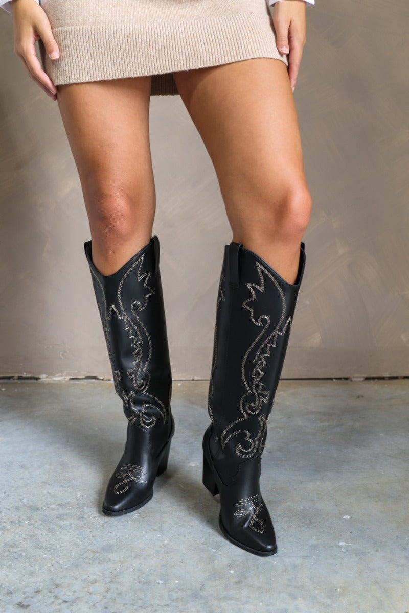 Front view of model wearing the Arcadia Black Western Boots that feature black faux leather, cream western stitching, and black soles with 3 inch heels.