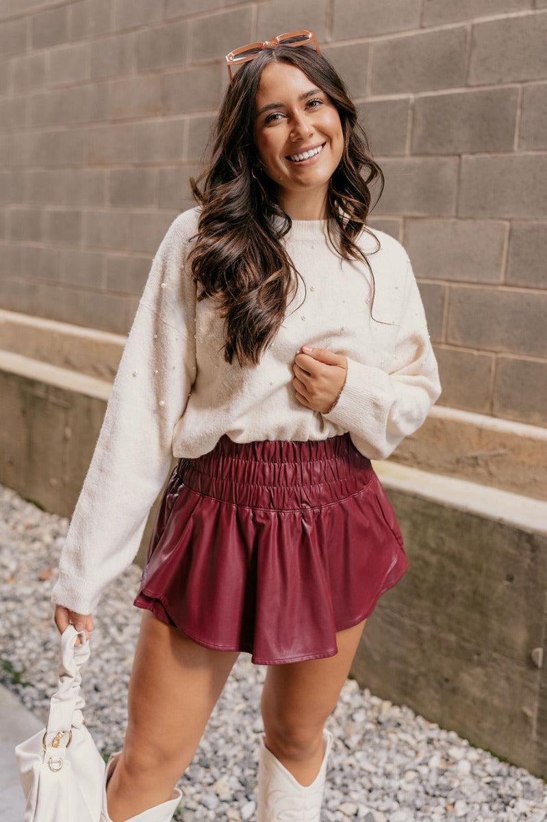 Front view of model wearing the Aurora Burgundy Faux Leather Ruffle Skort which features burgundy faux leather fabric, shorts lining, ruffle skirt detail, and a smocked elastic high waistband.