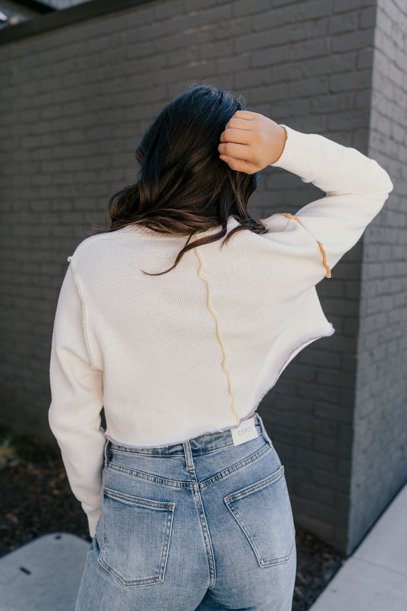 Back view of model wearing the Raegan Ivory Multi-Color Seam Sweater features white knit fabric with colorful exposed seam details, a high round neck, a cropped waist, and long sleeves.