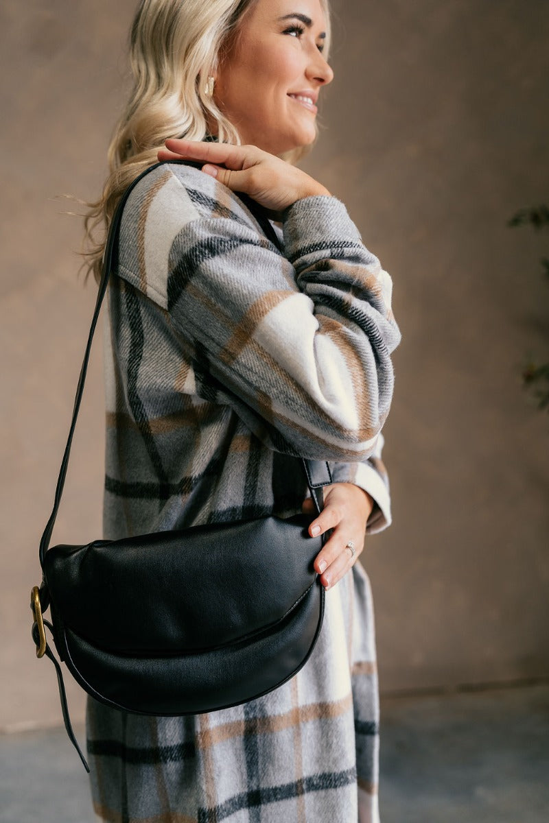 Front view of model wearing the Zoe Black Faux Leather Purse that has black faux leather fabric, an adjustable strap with a gold buckle detail, a snap button closure, and pockets on the inside.