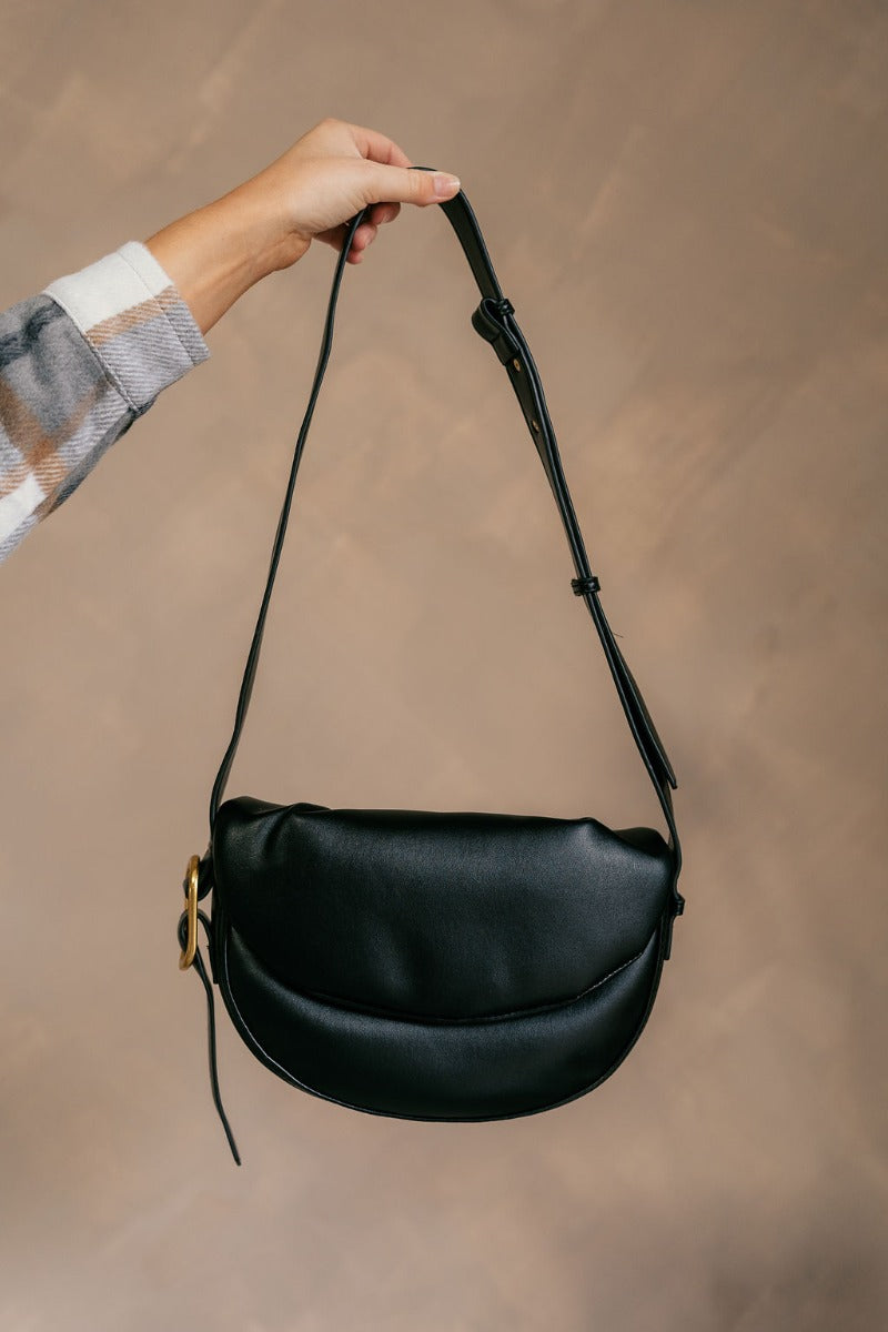 Model is holding the Zoe Black Faux Leather Purse that has black faux leather fabric, an adjustable strap with a gold buckle detail, a snap button closure, and pockets on the inside.
