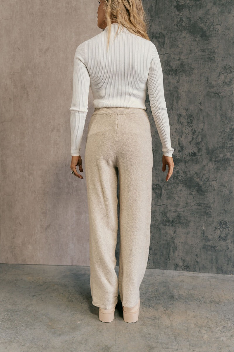 back view of model wearing the Destiny Oatmeal Knit Pants that have oatmeal knit fabric, a ribbed elastic waistband, and wide legs. 