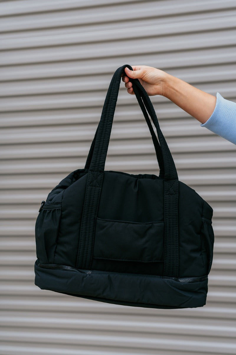 Front view of model holding the Isla Black Tote Bag that has black lightweight fabric, pockets on each side, an inside divider with a monochrome zipper, and shoulder straps.