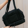 Front view of model holding the Isla Black Tote Bag that has black lightweight fabric, pockets on each side, an inside divider with a monochrome zipper, and shoulder straps.