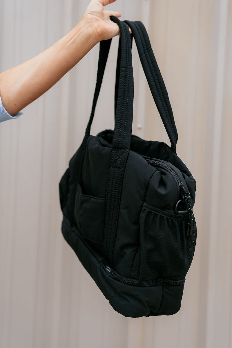 Side view of model holding the Isla Black Tote Bag that has black lightweight fabric, pockets on each side, an inside divider with a monochrome zipper, and shoulder straps.