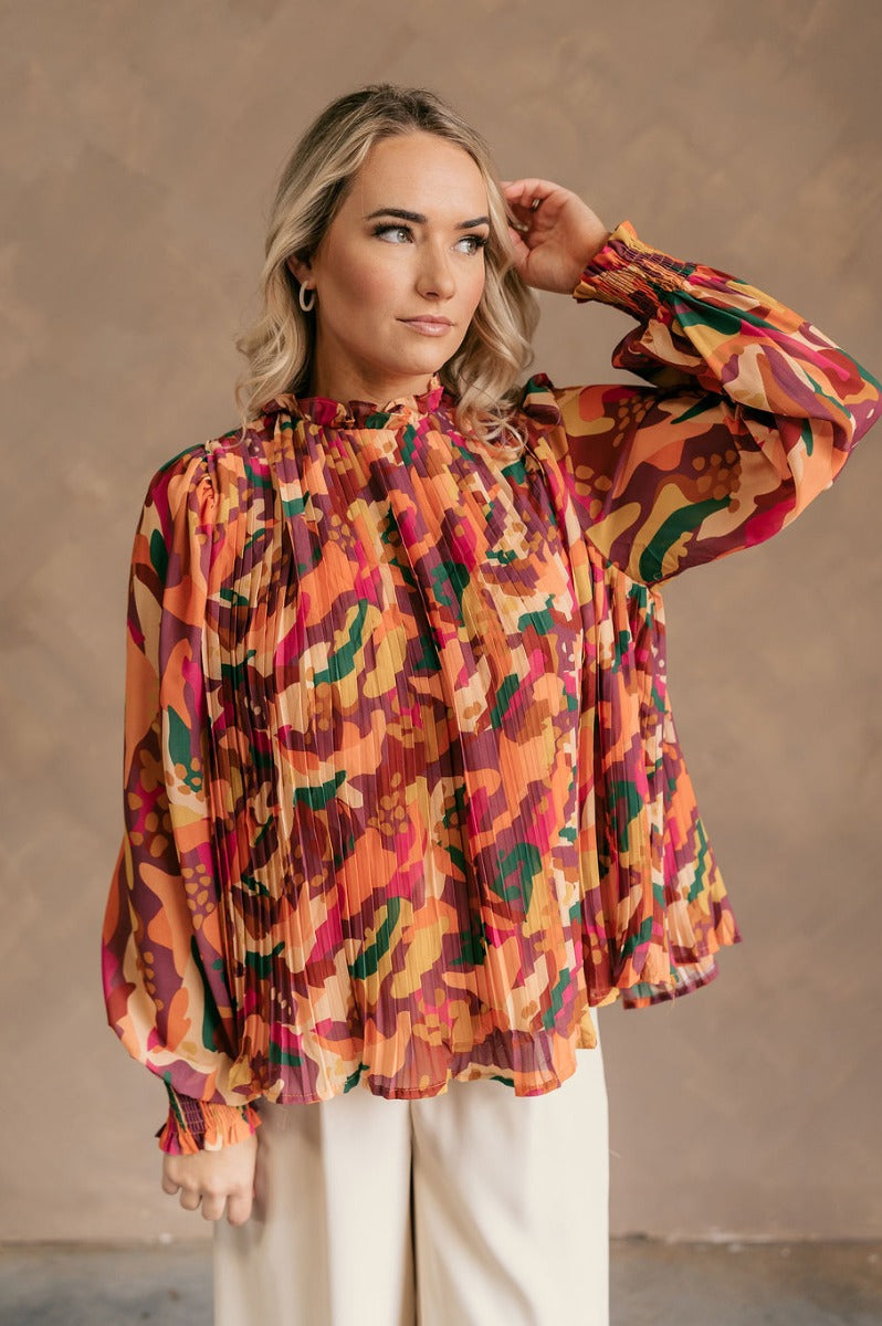 Front view of model wearing the Wrenley Multi Long Sleeve Top which features brown, orange, pink, green, purple, mustard, fuchsia and tan sheer fabric, pleated fabric, geomertic design, tan lining, high neckline with ruffle details, back key hole with but