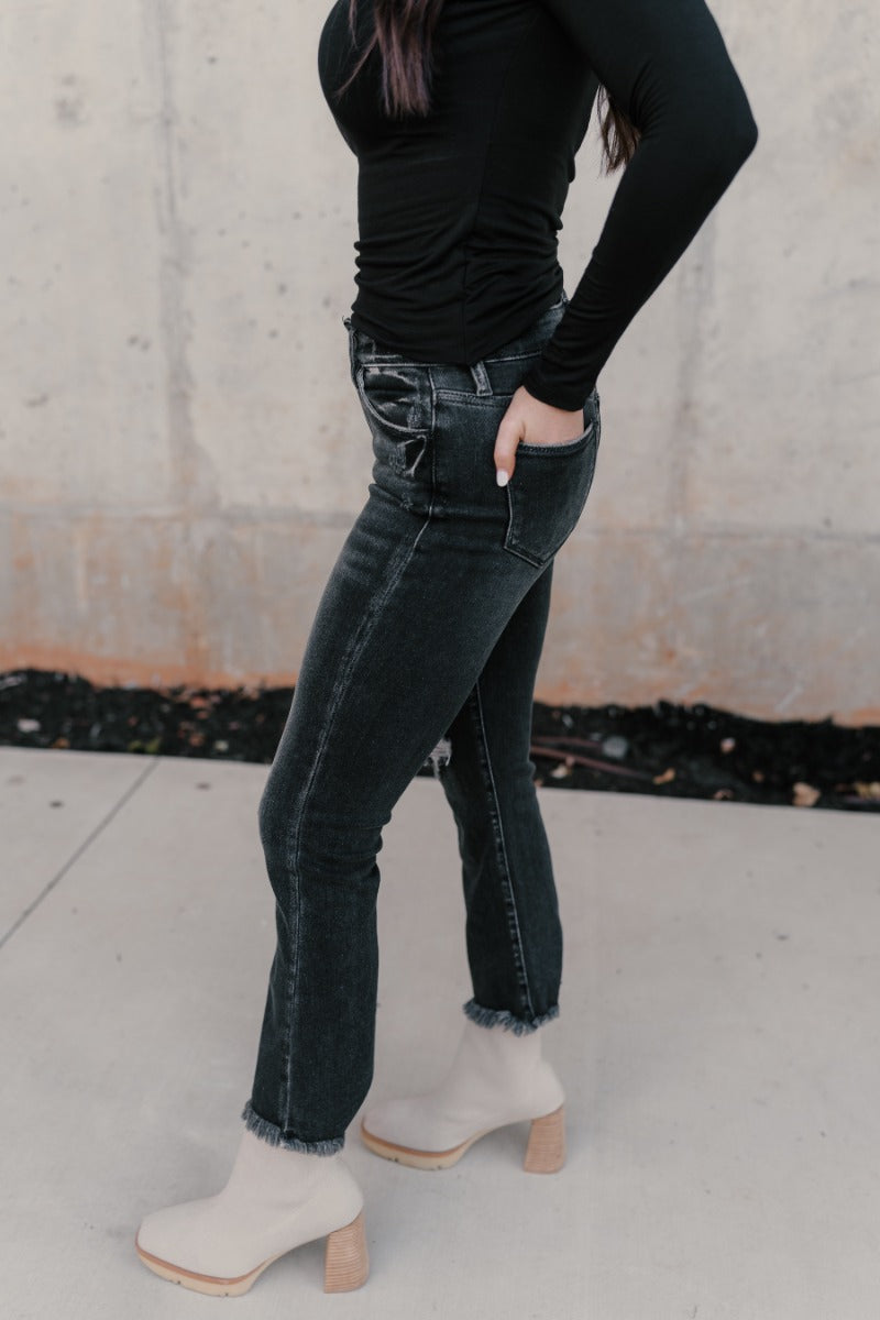 Side view of model wearing the Rooted Denim: Sydney Black Distressed Jeans that have distressed details, a front zipper belt loops, pockets and cropped flare legs with fray hem.