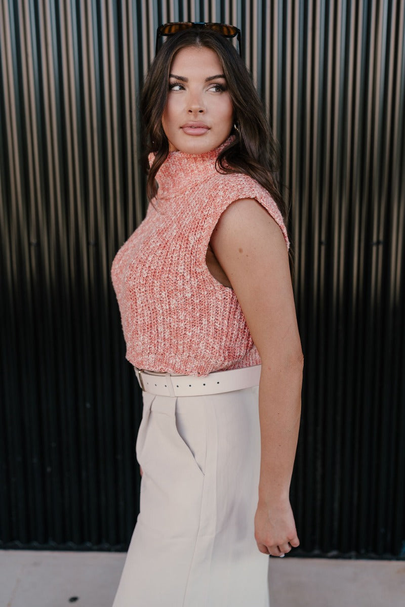 Side view of model wearing the Jordyn Orange & White Sleeveless Turtleneck Sweater which features orange and white cable knit fabric, a cropped waist, a turtleneck neckline, and a sleeveless design.
