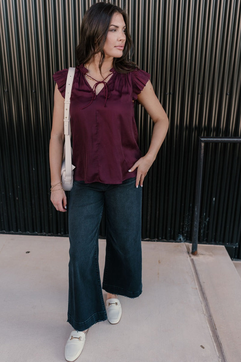 Full body view of model wearing the Angela Plum Satin Ruffle Short Sleeve Top which features plum satin fabric, a v-neckline with ruffle details, an adjustable tie, and short sleeves with pleated details.