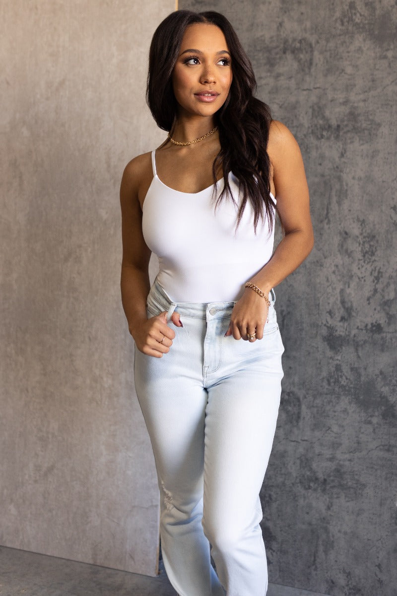 Front view of model wearing the Vera White Sleeveless Bodysuit which features white knit fabric, v-neckline, adjustable spaghetti straps, sleeveless and thong bottom with button snap closure.