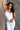 Front view of model wearing the Vera White Sleeveless Bodysuit which features white knit fabric, v-neckline, adjustable spaghetti straps, sleeveless and thong bottom with button snap closure.