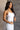 Side view of model wearing the Vera White Sleeveless Bodysuit which features white knit fabric, v-neckline, adjustable spaghetti straps, sleeveless and thong bottom with button snap closure.