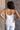 Back view of model wearing the Vera White Sleeveless Bodysuit which features white knit fabric, v-neckline, adjustable spaghetti straps, sleeveless and thong bottom with button snap closure.