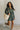 Full body front view of model wearing the Ezra Grey Green Long Sleeve Romper that has grey green knit fabric, pockets, an elastic waist, a front zipper, a hood, a skirt with fitted shorts , and long sleeves.