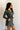 Side view of model wearing the Ezra Grey Green Long Sleeve Romper that has grey green knit fabric, pockets, an elastic waist, a front zipper, a hood, a skirt with fitted shorts , and long sleeves.