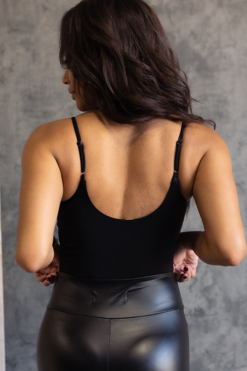 Back view of model wearing the Vera Black Sleeveless Bodysuit which features black knit fabric, v-neckline, adjustable spaghetti straps, sleeveless and thong bottom with button snap closure.
