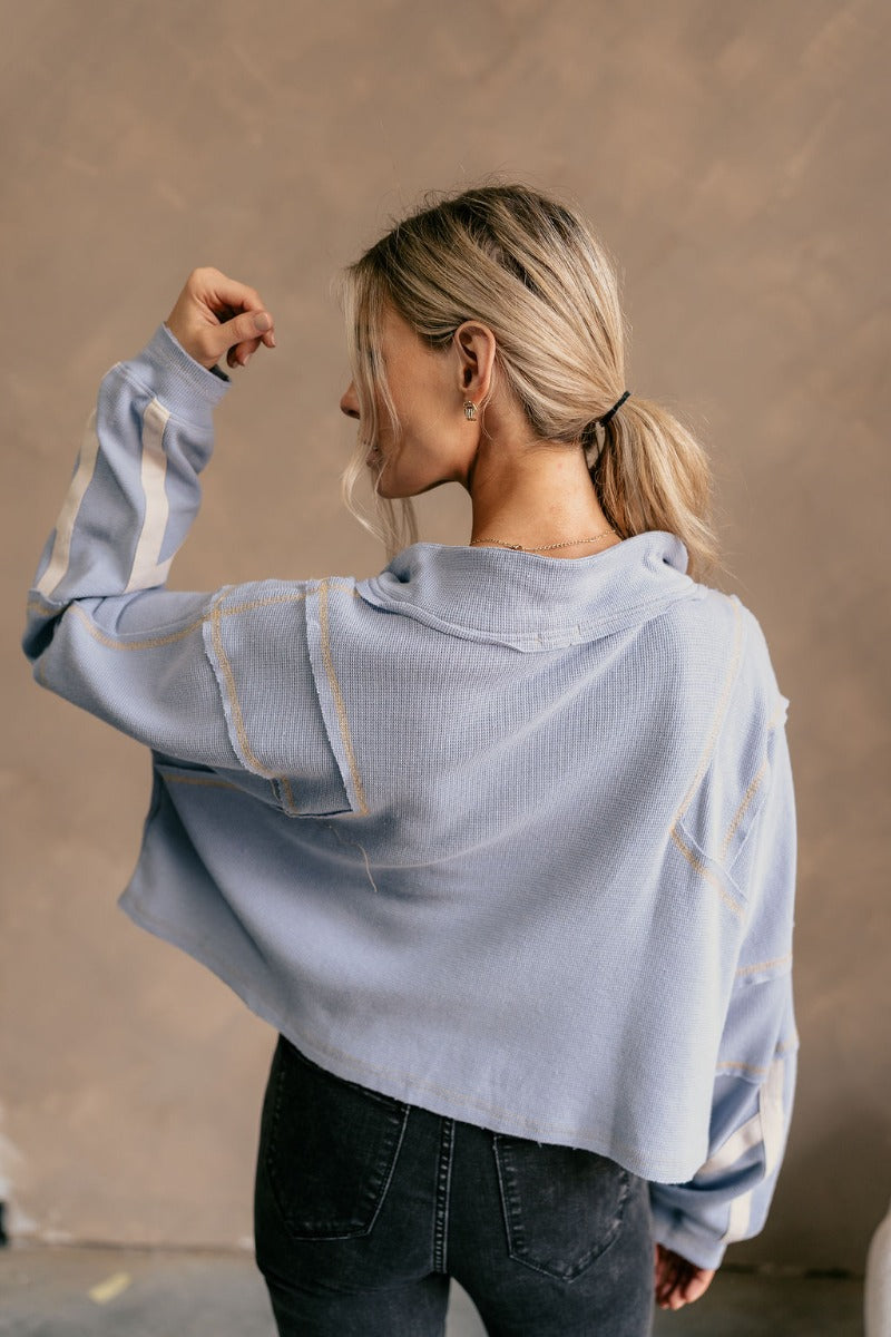 Back view of model wearing The Anna Periwinkle Thermal Long Sleeve Top features periwinkle blue knit fabric, light mustard threading, cream stripe design on the sleeves, cropped waist, one front pocket, quarter zip up, high neckline, distressed details an