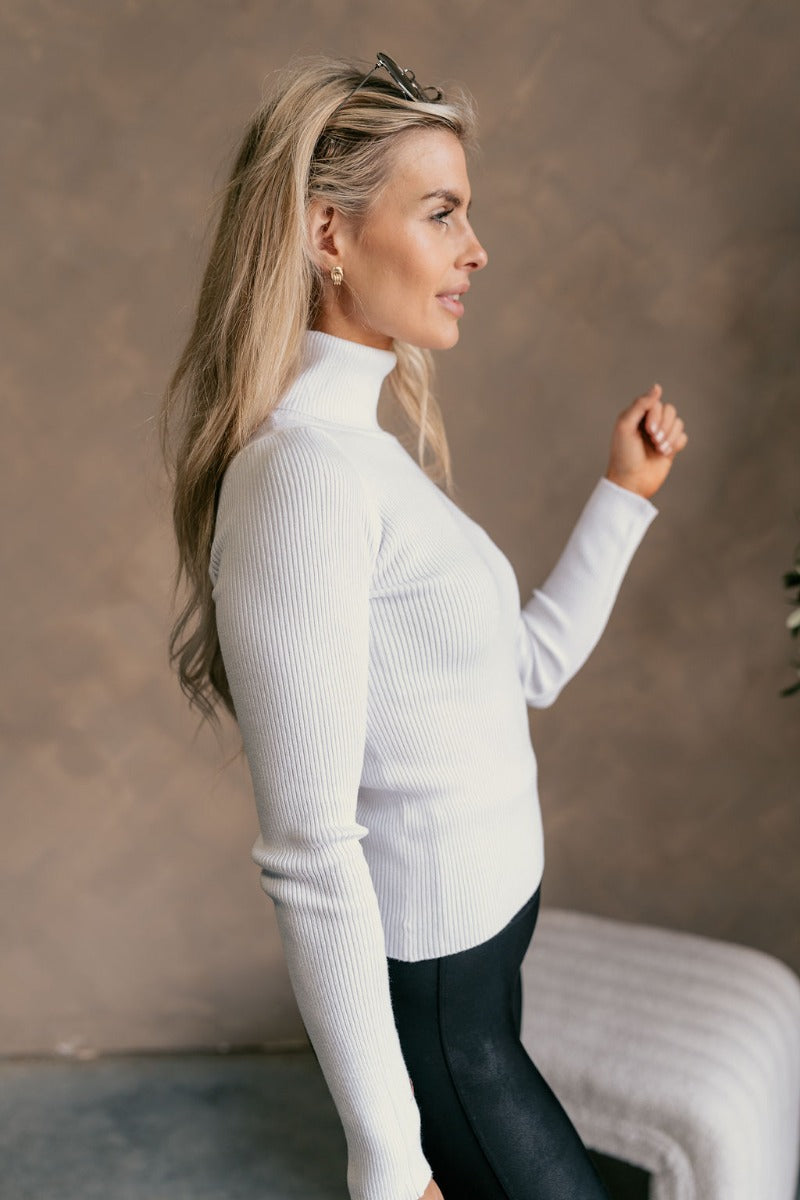 Side view of a model wearing The Elora Off White Long Sleeve Ribbed Turtleneck features off white ribbed fabric, turtleneck neckline and long sleeves.