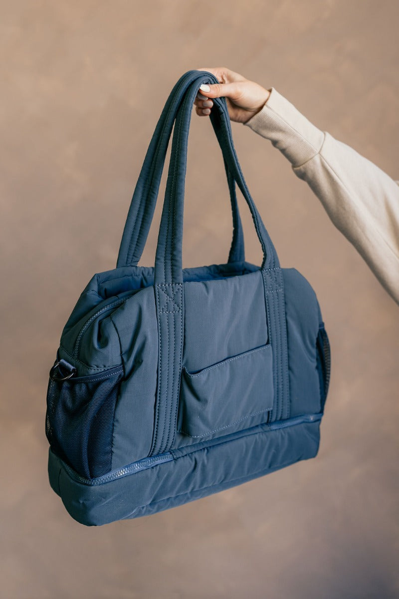 Front view of model holding the Isla Blue Tote Bag which features teal lightweight fabric, quilted pattern, pockets on each side, inside divider with monochrome zipper and shoulder straps.