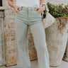 Front view of model wearing the Livia Sage Wide Leg Pants that has sage green washed denim-like fabric, a front zipper with a button closure, belt loops, front pockets, back pockets and wide pant legs.