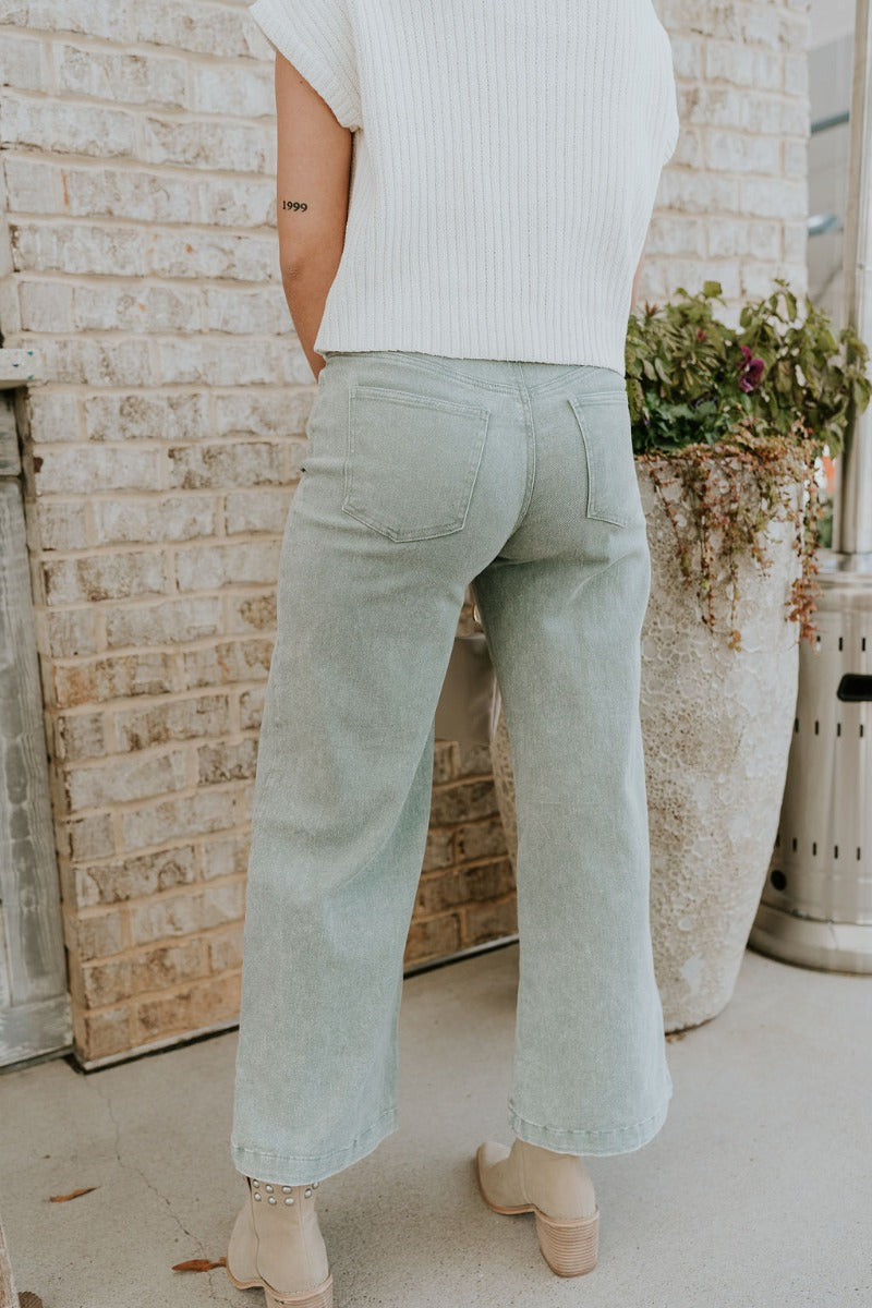 Back view of model wearing the Livia Sage Wide Leg Pants that has sage green washed denim-like fabric, a front zipper with a button closure, belt loops, front pockets, back pockets and wide pant legs.