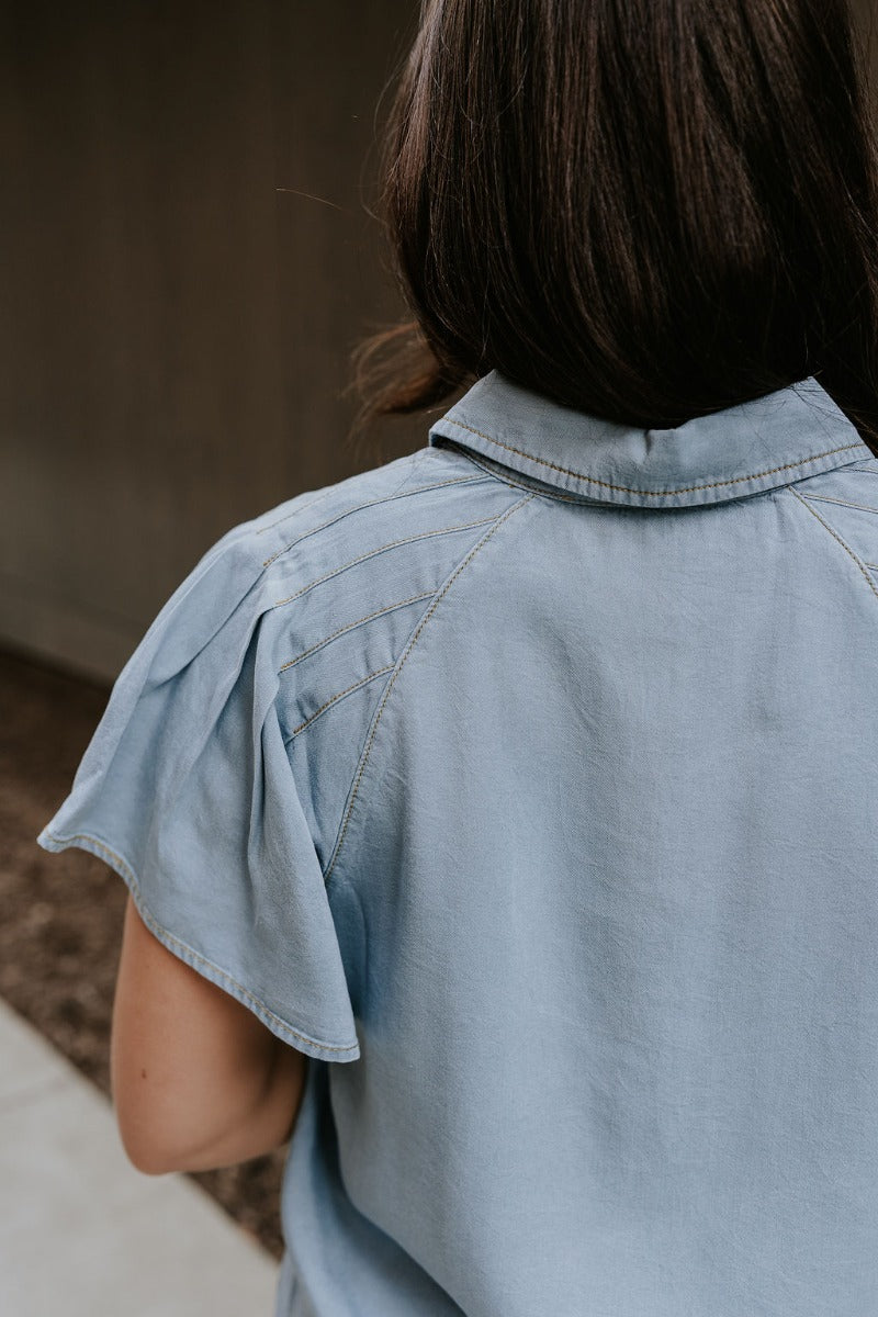 Upper back view of model wearing the Cynthia Blue Short Sleeve Button Up Top that has light blue tencel fabric, light brown stitching , grey tortoise buttons, a collared neckline, and short flare sleeves.